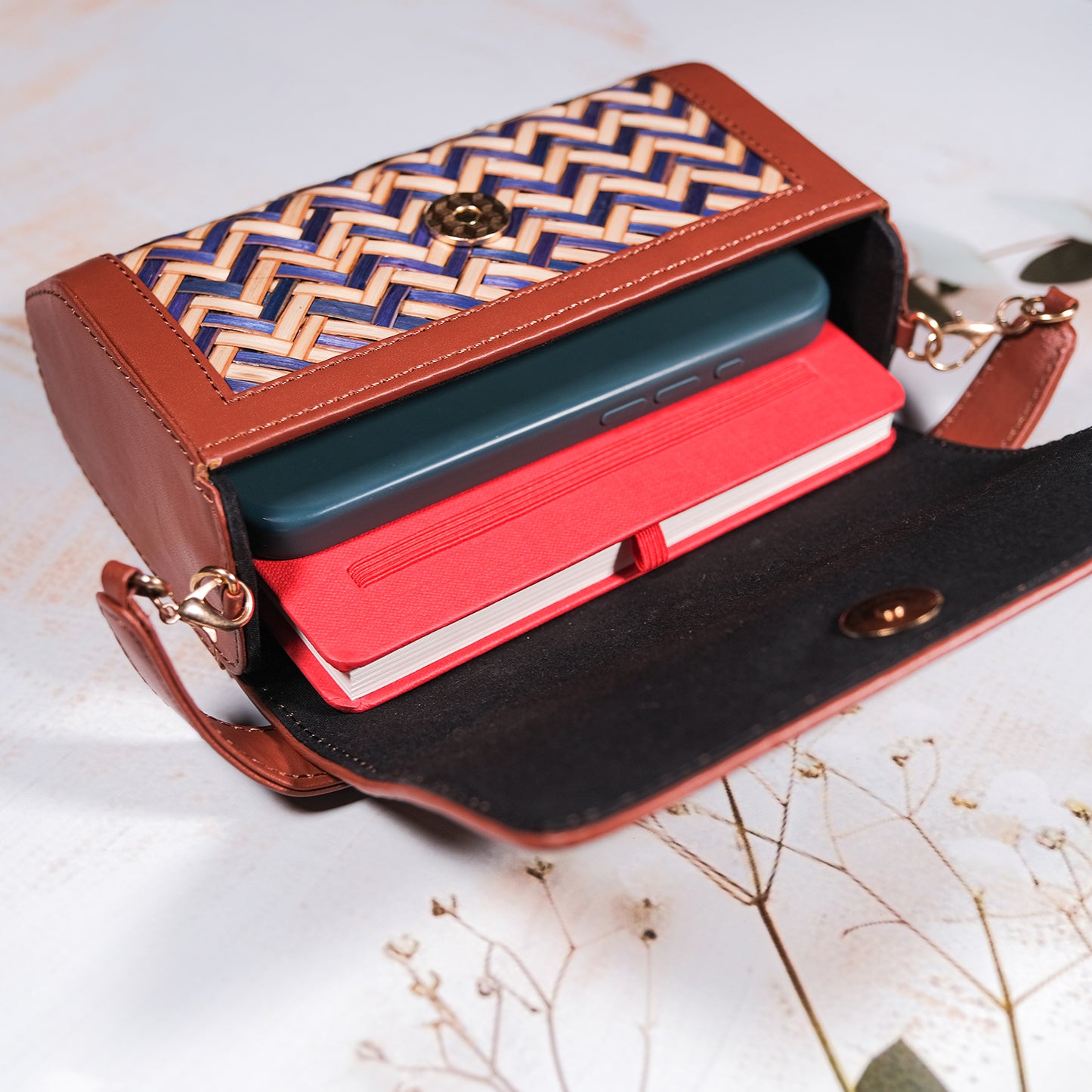 Open brown handbag with a patterned interior, containing a red notebook and a green phone, on a light background.