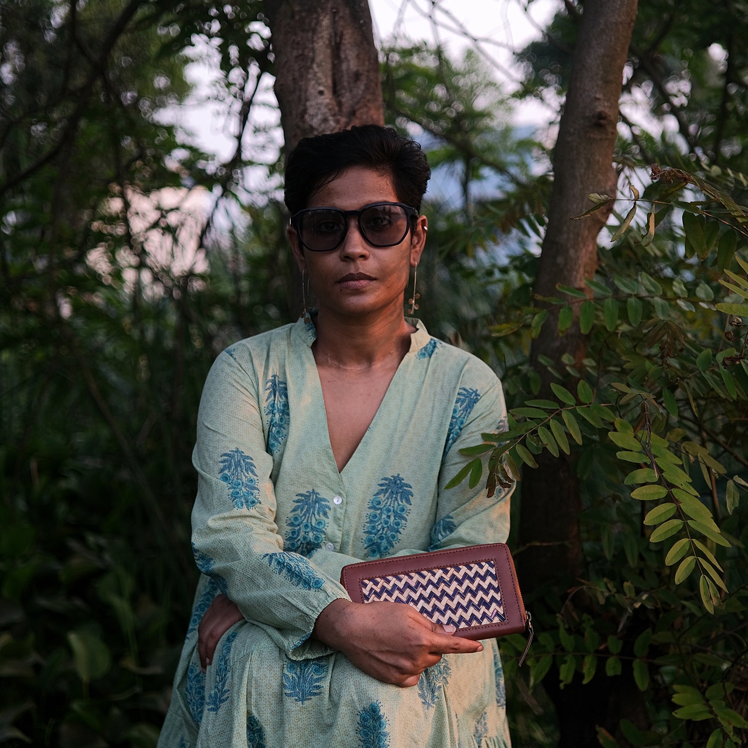 Person wearing sunglasses and a light-colored outfit with a pattern, holding a clutch in a forest setting.