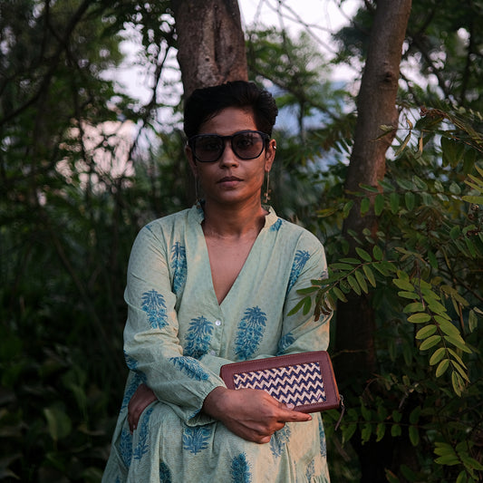 Person wearing sunglasses and a light-colored outfit with a pattern, holding a clutch in a forest setting.