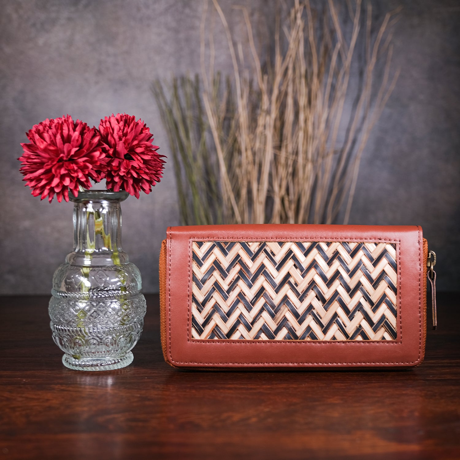 Brown clutch bag with chevron pattern next to a vase with red flowers on a wooden surface.