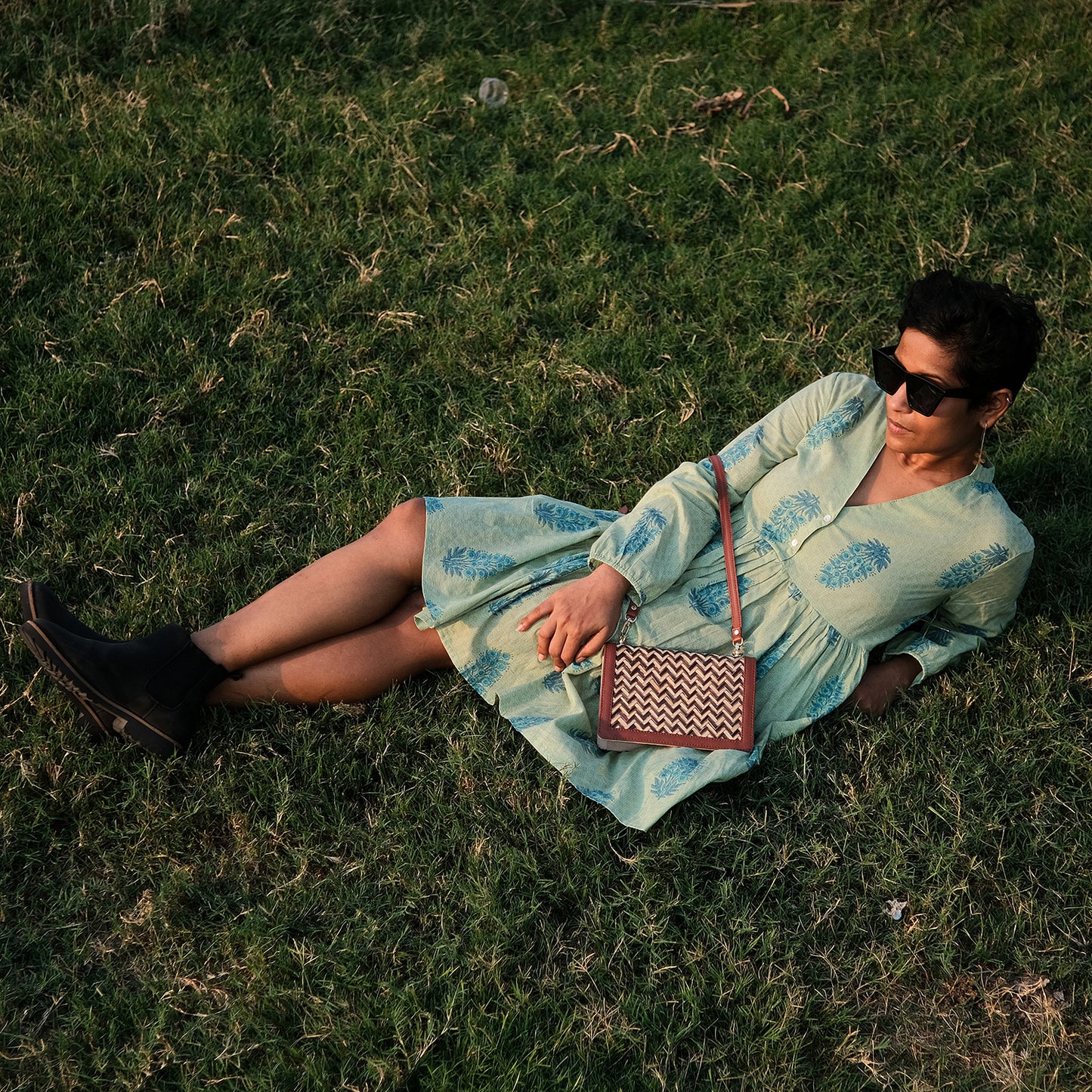 Woman in a light blue dress sitting on grass with a patterned handbag.