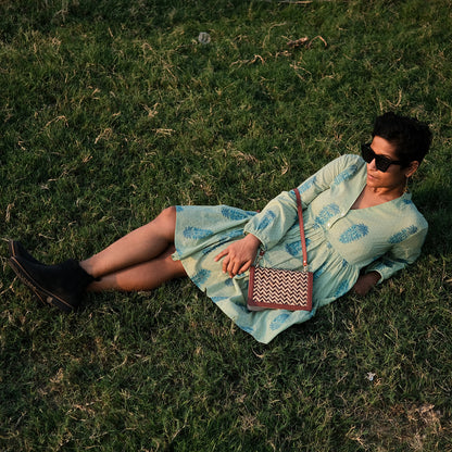 Woman in a light blue dress sitting on grass with a patterned handbag.