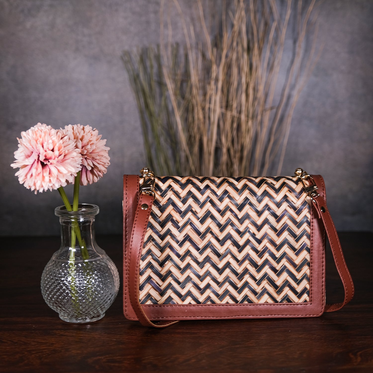 Woven handbag with zigzag pattern next to a vase with pink flowers on a dark surface.