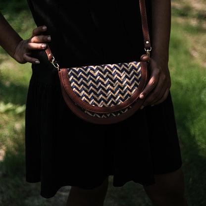 A person holding a half moon shaped sling bag with a chevron weave pattern in blue and beige colors and brown leather accents.