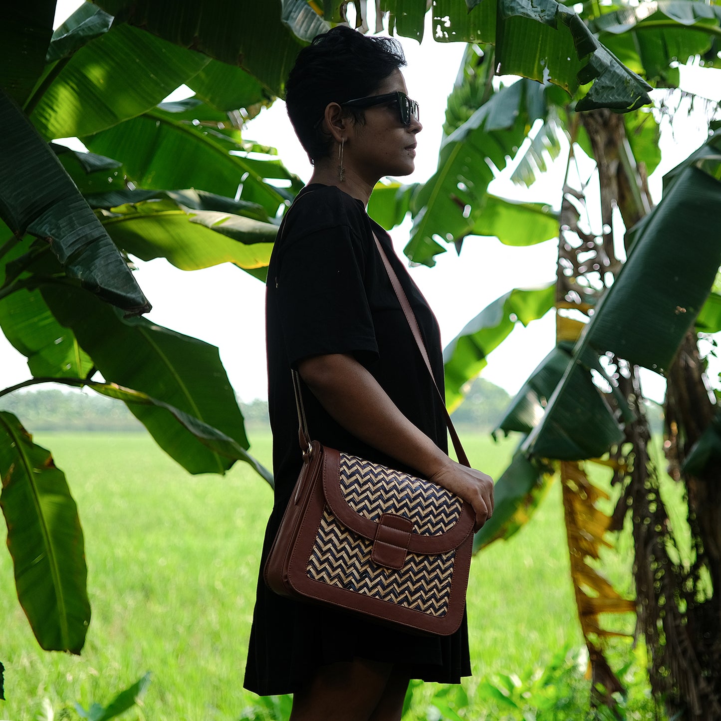 A person standing in a natural setting with green foliage in the background, wearing a black dress and carrying a brown and blue zigzag patterned sling bag.