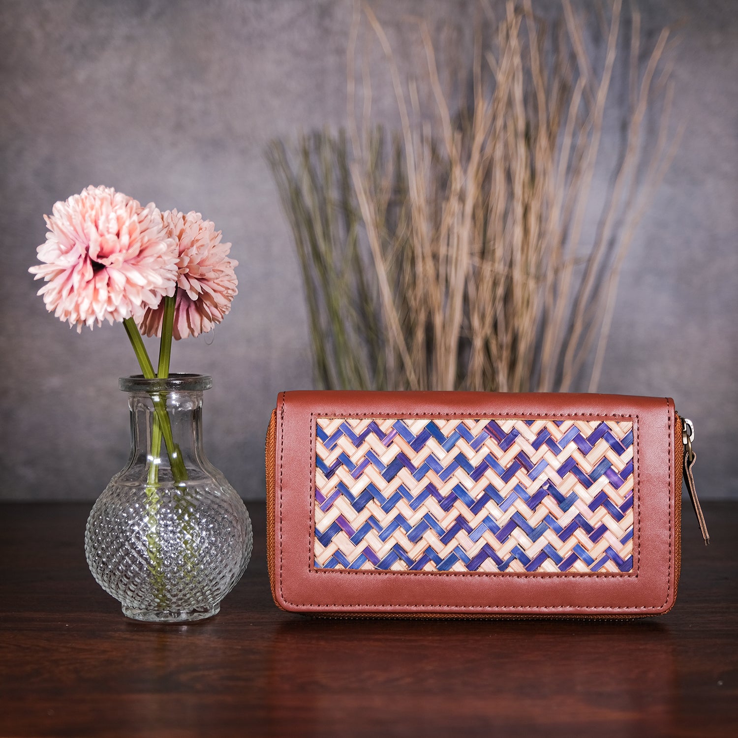Brown clutch with wicker pattern next to a vase with pink flowers and a plant on a wooden surface.
