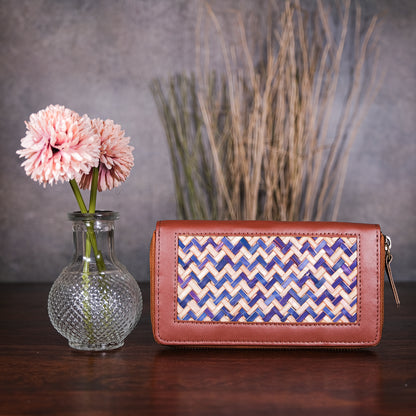 Brown clutch with wicker pattern next to a vase with pink flowers and a plant on a wooden surface.