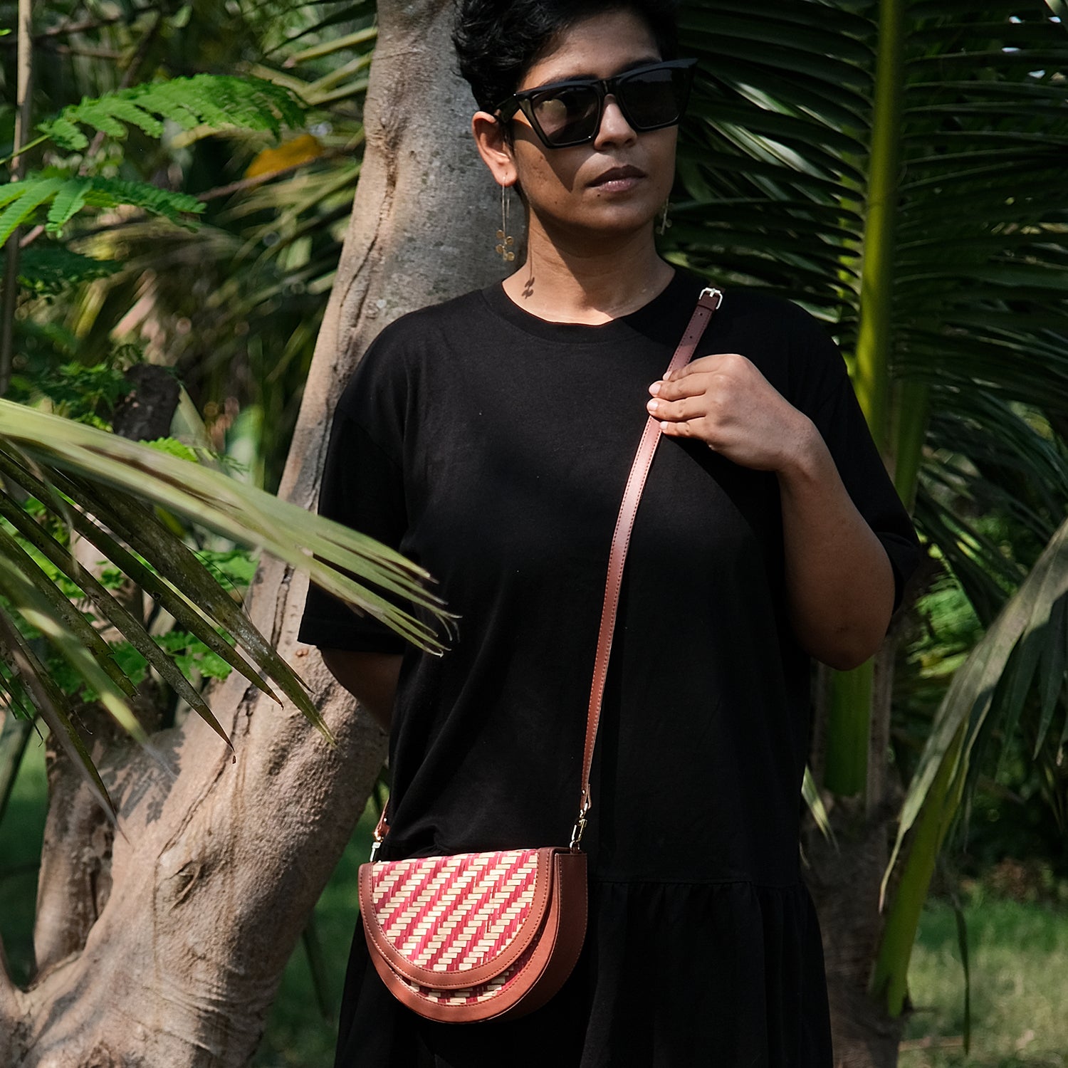 A person wearing sunglasses and a black dress is standing in a tropical setting with palm trees in the background, carrying a half-moon shaped sling bag with a red and beige chevron pattern and brown leather accents.